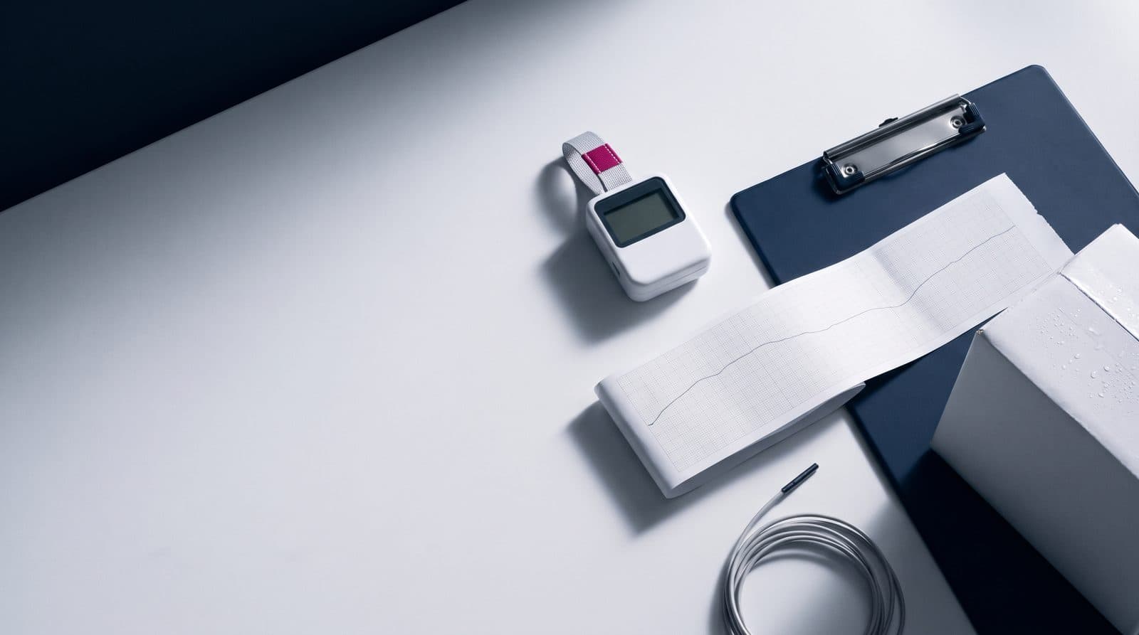 Overhead still life: a temperature logger with a strap, a paper strip with a temperature curve, a navy clipboard and a white carton with condensation on a light desk.