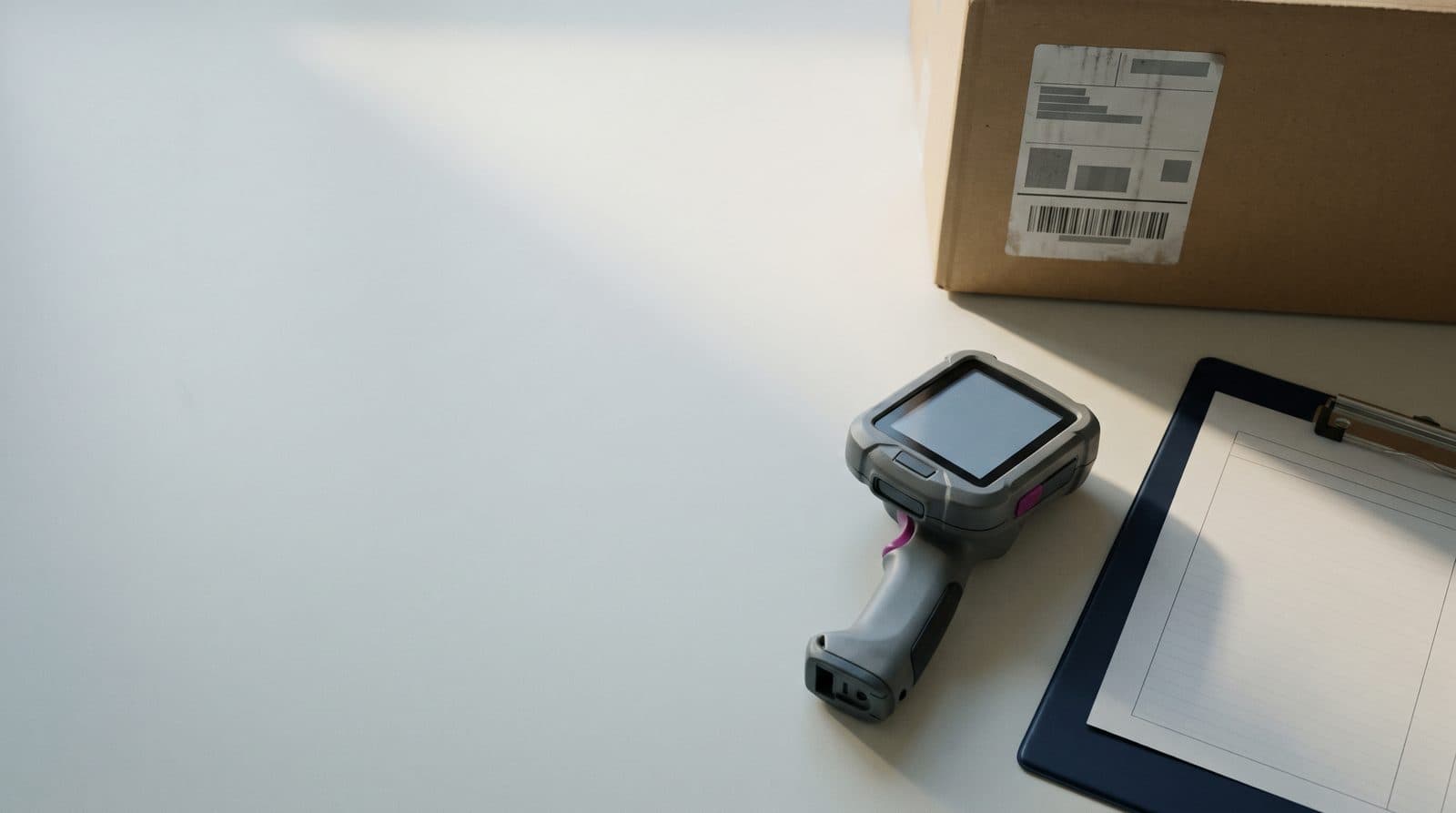 Overhead still life: the corner of a carton with its original supplier label, a handheld scanner and a clipboard with a blank sheet.