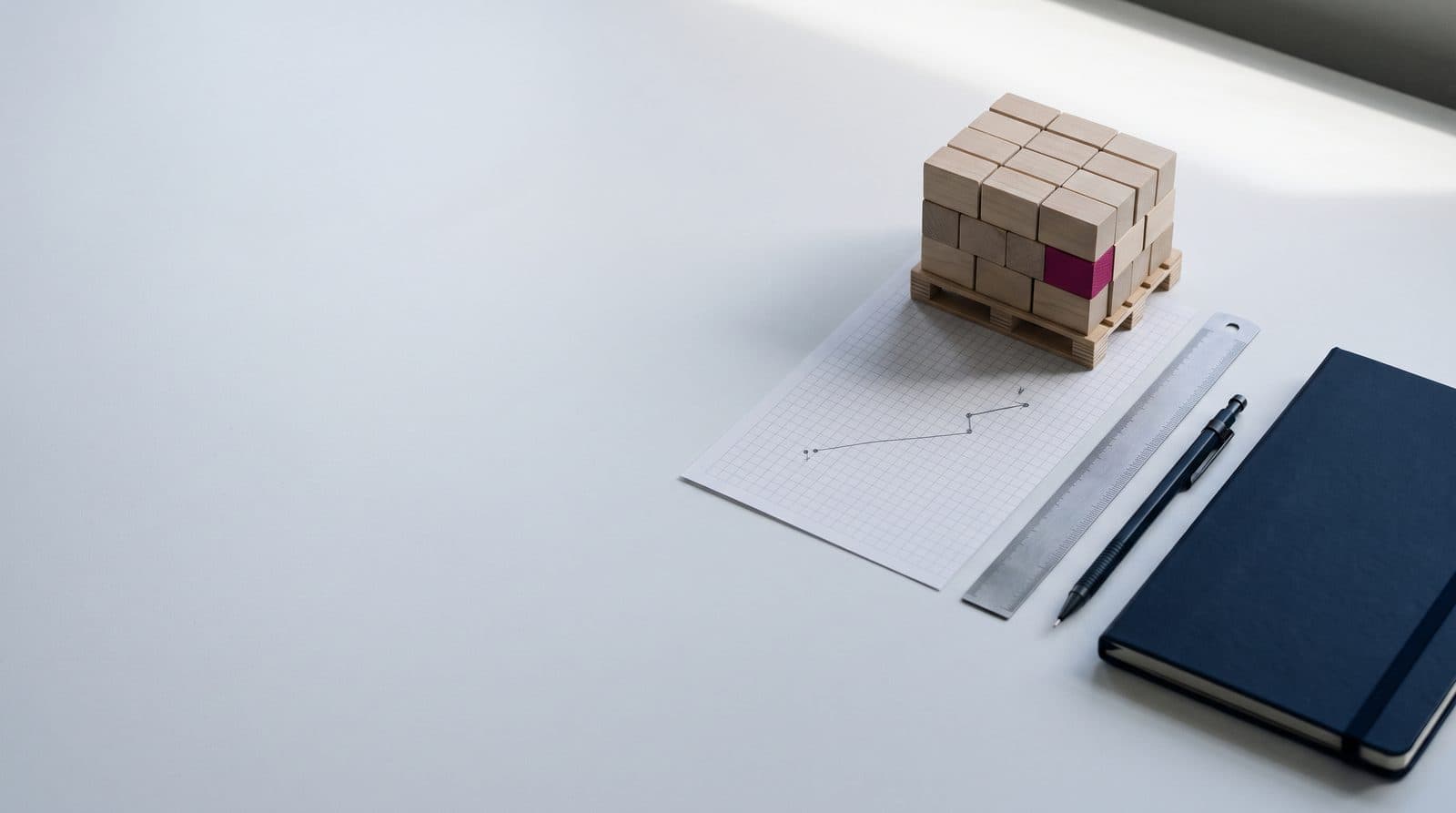 Overhead still life: a scale pallet with wooden blocks, grid paper with a pencil route, a ruler, a mechanical pencil and a navy notebook.