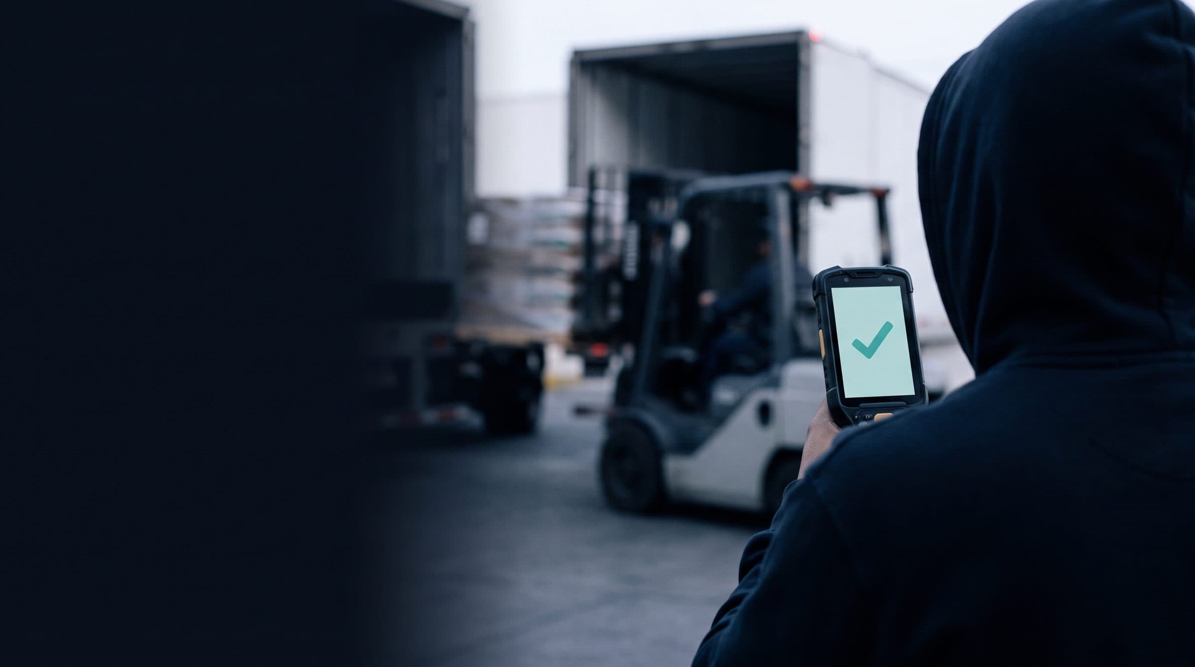 A hooded operator, seen from behind, holds a scanner showing a check mark while a forklift loads a trailer.