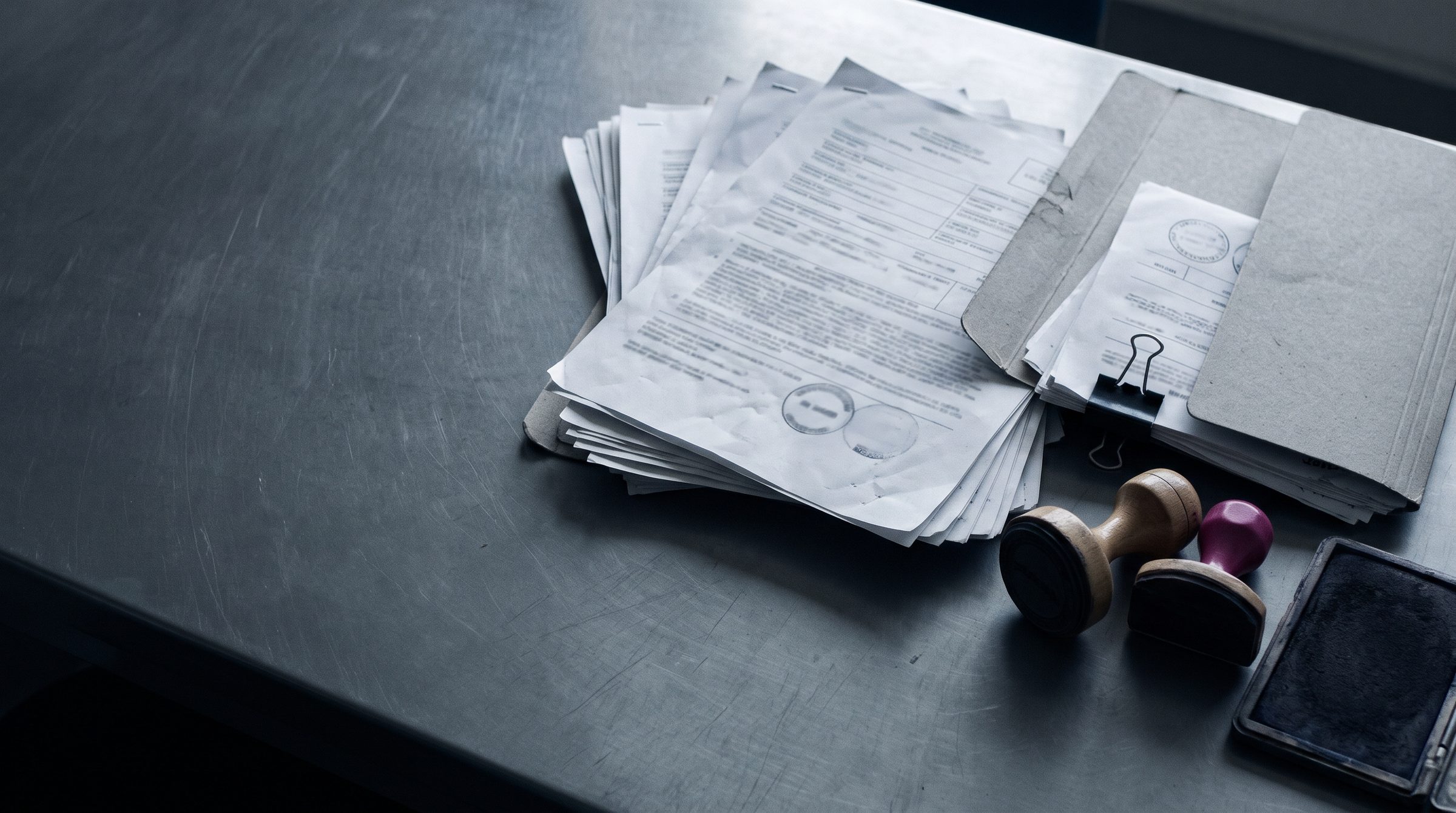 Sobre un escritorio metálico, una pila de documentos de embarque y aduana, una carpeta con clip y dos sellos de goma.