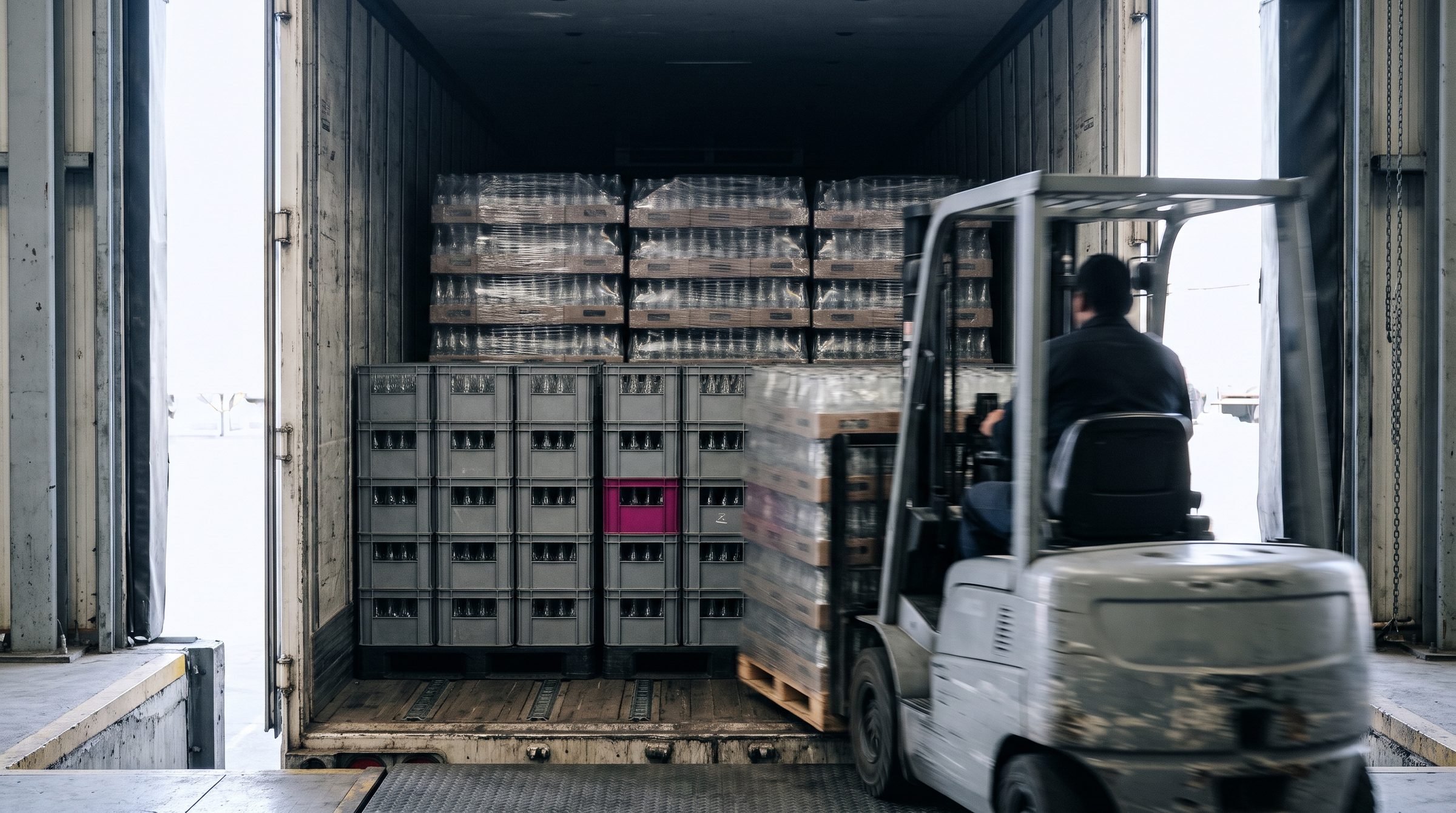 Interior de un tráiler cargado con camadas de envases retornables de vidrio en la base y paquetes emplayados encima; un montacargas entra el siguiente palet.